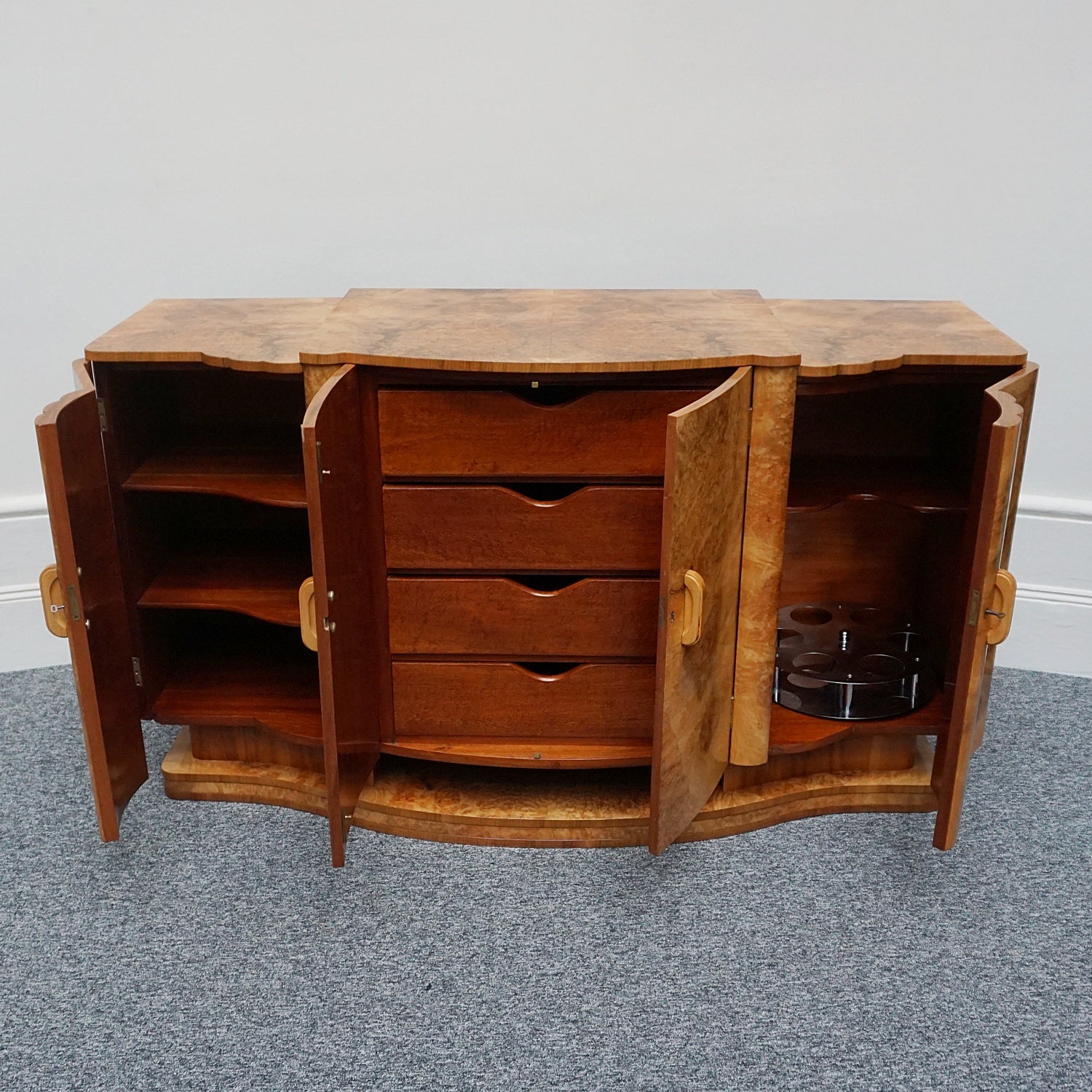 An Art Deco Sideboard by Harry & Lou Epstein with fluted sides set over a curved walnut base made of solid mahogany with burr walnut veneer. Left compartment contains a stainless steel rotating bottle holder with upper mahogany shelf. The central section contains four large mahogany lined drawers, with the left hand side containing shelves.