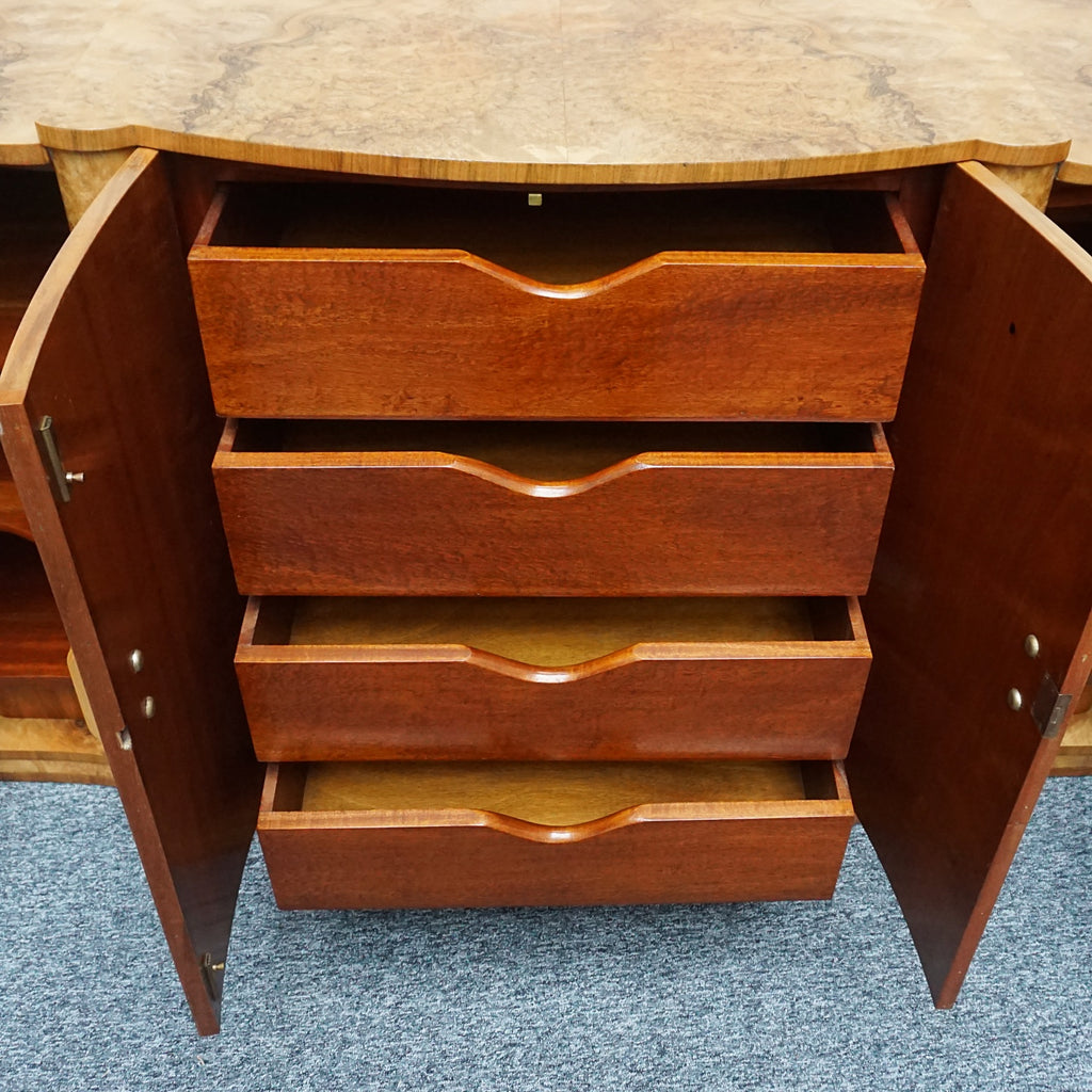 An Art Deco Sideboard by Harry & Lou Epstein with fluted sides set over a curved walnut base made of solid mahogany with burr walnut veneer. Left compartment contains a stainless steel rotating bottle holder with upper mahogany shelf. The central section contains four large mahogany lined drawers, with the left hand side containing shelves. 