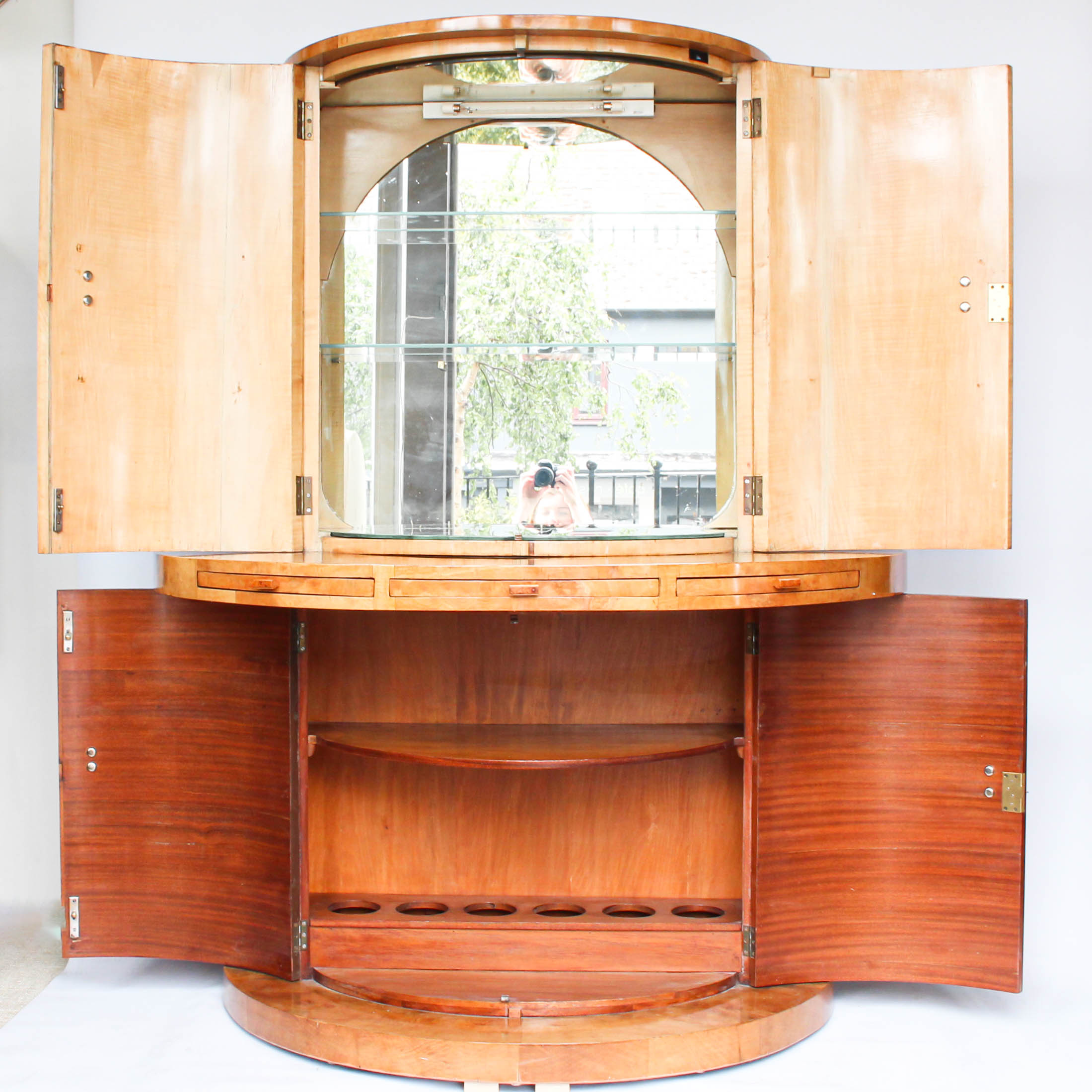 An Art Deco, demi-lune cocktail cabinet attributed to Harry & Lou Epstein. Mirrored, lit interior to top with glass shelves and mirrored slide tray. Lower, shelved cabinet with bottle holders. Burr walnut veneer at Jeroen Markies.