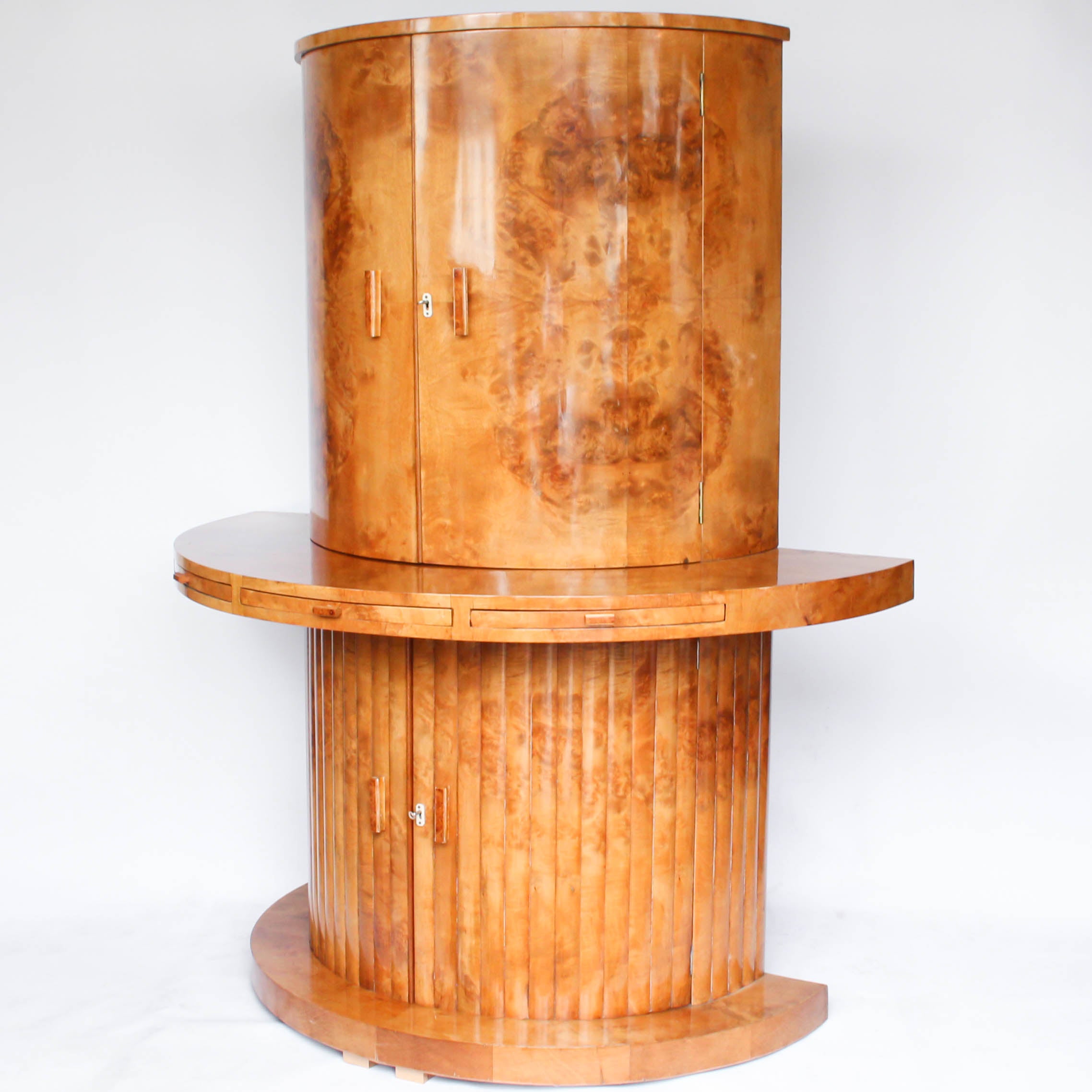 An Art Deco, demi-lune cocktail cabinet attributed to Harry & Lou Epstein. Mirrored, lit interior to top with glass shelves and mirrored slide tray. Lower, shelved cabinet with bottle holders. Burr walnut veneer at Jeroen Markies.